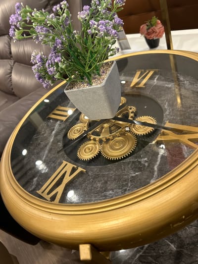 The image shows a close-up of a decorative table clock with Roman numerals and visible gold gears. A small potted plant with purple flowers sits on top of the clock. The clock has a black marble-patterned face and a gold frame. In the background, a brown leather couch and a small pink artificial flower arrangement on a white surface are visible. The scene appears to be indoors, possibly in a home or commercial setting. The location in Makassar, Indonesia, is not discernible from the image itself.