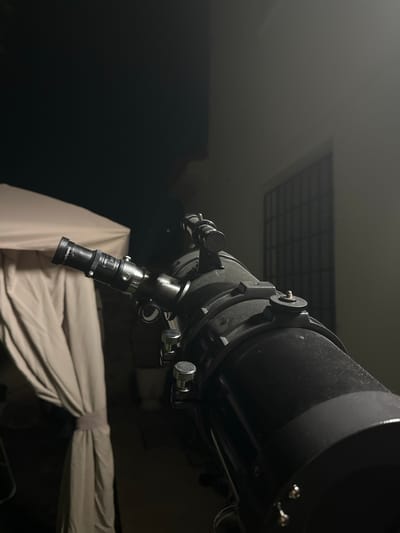 A nighttime outdoor scene in Villa Pasquali, Italy, features a dark-colored astronomical telescope positioned prominently in the foreground, aimed upwards and slightly left. The telescope is a large reflector type, dark grey or black, with visible dust or specks on its surface. Attached is a black eyepiece marked "Eyepiece" and "5X Lens", along with a smaller black finder scope mounted parallel to the main tube. Silver-colored adjustment knobs and fasteners are present on the telescope body.

To the left, a light-colored, possibly beige or off-white, fabric canopy or tent structure is partially visible, with one side draped and tied back. To the right, a building wall, also light-colored, is illuminated, featuring a dark, grid-patterned window. The sky above is dark, indicating night. No individuals are present in the image.