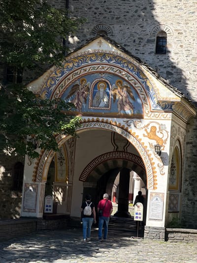 The image depicts the ornate entrance to the Rila Monastery in Rilski Monastery, Bulgaria. The entrance is adorned with vibrant frescoes, featuring religious figures like angels and a central portrait of a saint, surrounded by intricate floral and geometric patterns in shades of blue, yellow, and white. A carved lion is visible on the right side of the entrance arch. Two people, a man wearing a red shirt and jeans and a woman in a white backpack and blue jeans, are walking away from the camera towards the entrance. A third silhouetted figure is visible in the background through the archway. The ground is paved with cobblestones, and a leafy tree branch extends into the frame from the left, casting shadows. The architecture is characterized by rough stone walls and arched passageways. Information boards are mounted on the walls near the entrance.