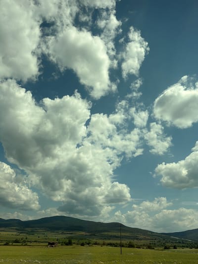 This media file captures a landscape scene, likely in Mirovo, Bulgaria. The image is dominated by a vast expanse of blue sky filled with fluffy white cumulus clouds. The clouds are varied in size and density, with some appearing wispy and others more substantial, suggesting a partly cloudy day.

The lower portion of the image reveals a verdant landscape. A gently sloping green field, dotted with small white wildflowers, occupies the right side. Beyond the field, a dark, heavily wooded mountain range stretches across the frame, its peak shrouded in shadow and mist. The mountainside shows signs of human activity, with agricultural fields and scattered trees visible in the middle ground. A single, dark utility pole stands prominently in the foreground of the field, and a small, rustic-looking building or shed is nestled into the base of the mountain. The presence of cultivated land and the mountain range suggest a rural setting.

The lighting indicates it is daytime, likely late morning or early afternoon, given the bright sunlight illuminating the clouds and the field. The weather appears pleasant, with a mix of sun and clouds, creating a dynamic sky. No people or specific events are depicted; the focus is on the natural scenery. There is no visible text in the image.