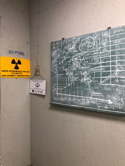 The image shows a corner of a room in Agia Paraskevi, Greece, where a whiteboard is mounted on the wall. The whiteboard is green and is covered in white chalk markings. These markings include numbers, symbols, a line graph, and some Greek text. The numbers and symbols appear to represent some kind of data. A radiation warning sign with a yellow background and a black radiation symbol is attached to the wall to the left of the whiteboard. The sign is in Greek and English and reads "HIGH INTENSITY RADIATION AREA." Below the sign is a smaller sign with a blue background and white text. The walls are painted a light gray. There are no people visible in the image. The room appears to be a laboratory or a workspace.