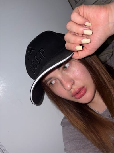 A woman with light skin and brown hair, wearing a grey shirt, is lying on a light surface, possibly a bed or table. She is wearing a black baseball cap with white trim and Cyrillic lettering. Her nails are painted a pale yellow color. She has a nose ring and is looking at the camera with her mouth slightly open. The location is not identifiable as being in the city of Port-Katon, Russia.