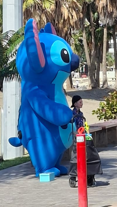 The image displays an outdoor scene in Los Cristianos, Spain, characterized by bright, sunny conditions. A large, inflatable blue costume, depicting the Disney character Stitch, stands on a light-colored paved surface, facing slightly to the right. The costume features black eyes with white pupils, pink inner ear details, and lighter blue markings on its chest and face. To the immediate right of the costume, a young child is positioned, looking towards the right side of the frame. The child wears a dark baseball cap, reversed, and a blue patterned shirt. The child's left hand is near the handle of a black wheeled cart, which is partially obscured by a prominent red vertical bollard located on the far right. The cart holds several brightly colored items, including yellow and green containers. A small, light blue rectangular box is situated on the paved ground near the base of the Stitch costume. The background reveals a sandy area backed by numerous palm trees, behind which indistinct buildings are visible.