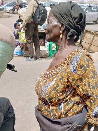 An outdoor scene in Masvingo, Zimbabwe, captures a busy public thoroughfare or market. In the right foreground, an elderly dark-skinned woman is depicted in profile, facing left. She wears a dark green headwrap, ornate gold-colored dangling earrings, and a brown and gold patterned dress adorned with black, white, and blue designs, featuring white beadwork along the neckline. A dark fabric is draped across her lower waist. In the midground, a male figure in a light brown shirt, dark trousers, and blue sandals stands, carrying a black backpack. Behind him, a woman in a blue headwrap is seated at a low table, actively engaged with items within a light green plastic basket, signifying a vending operation. A green plastic stool and wooden crates are positioned near the seated woman. The ground consists of an uneven, possibly unpaved or heavily worn asphalt surface. The background reveals multiple vehicles, primarily light-colored sedans, alongside several pedestrians and children, indicating a bustling urban environment.