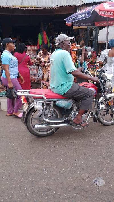 The image depicts a busy outdoor market scene in Agbarho, Nigeria. In the foreground, a man wearing a light blue t-shirt and grey trousers is riding a red motorcycle with a silver exhaust pipe and red seat. He is wearing a grey cap and black flip-flops. The motorcycle's rear wheel is prominent in the frame, with visible spokes and a silver fender. Behind the motorcycle, several people are browsing stalls. To the left, a woman in a light blue shirt and purple trousers holds a black plastic bag. Next to her, another woman in a red dress is visible. Further back, a woman in a patterned dress is interacting with someone at a stall laden with packaged goods. Above the stalls, a striped awning is visible, with a red and blue umbrella bearing text. The ground is paved with asphalt. The overall atmosphere suggests a lively commercial environment with diverse goods and activities.