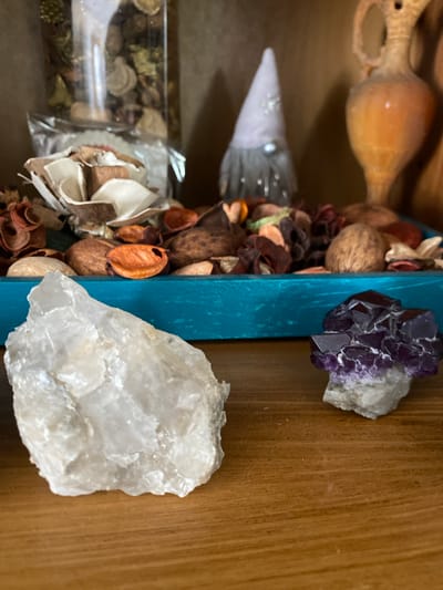 A still life composition features two rocks on a wooden shelf. The rock on the left is a large, translucent white quartz. The rock on the right is a smaller cluster of dark purple amethyst crystals. Behind them, a turquoise tray holds dried potpourri. In the background, a glass jar filled with dried botanicals and a small gnome figure are visible. To the far right, a terracotta-colored jug with handles sits on the shelf. The image is taken indoors, likely in a residential setting within Osek, Czechia.