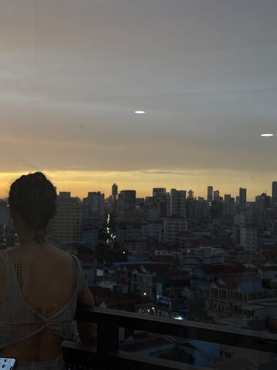 A woman stands on a balcony overlooking a city skyline at dusk. The woman is facing away from the camera, with her back to the viewer. She has dark hair pulled up in a bun and a tattoo on her neck. She is wearing a light-colored, open-back top and appears to be leaning on the balcony railing. The city below is densely populated with buildings of varying heights, and lights are beginning to illuminate the streets. The sky is a gradient of orange and grey, indicative of sunset or twilight. The image was taken in Khan Mean Chey, Cambodia.