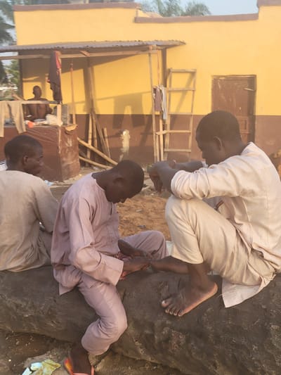 Three African males are present in an outdoor setting near a building in Kwamba, Nigeria. One man is crouched on a large, dark rock with bare feet, wearing a light-colored, long-sleeved outfit. A second man sits on the rock, clad in a pinkish-purple, long-sleeved ensemble. A third man is partially visible, kneeling and wearing a light-colored outfit. A building with a mustard-yellow exterior is in the background, featuring a boarded-up window and wooden scaffolding.