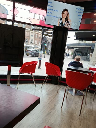 The image shows the interior of a commercial establishment with a large window overlooking a street. On a digital screen mounted above the window, an image of singer Tate McRae is displayed, accompanied by song titles. Visible outside the window are buildings, street signs, and people walking. Specifically, street signs indicate "Lurke Street," "The Higgins," and "Church Arcade," suggesting the location is in Bedford, United Kingdom. Red chairs are arranged within the establishment, and a person can be seen from behind sitting at a table. The flooring appears to be light-colored wood.