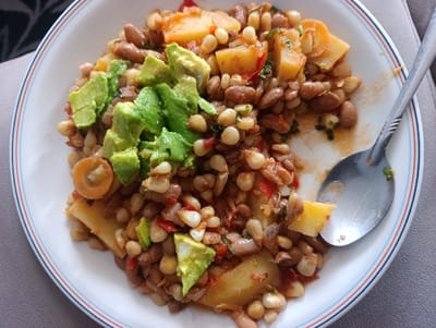 An overhead shot displays a partially consumed meal served in a white oval plate with a blue and red striped rim, resting on a light grey surface. The meal consists of a mixture of pale yellow maize kernels, light brown beans, diced yellow potatoes, chopped red bell pepper, and green herbs, all coated in a reddish-brown sauce. Several bright green diced avocado pieces are arranged on top of the food, primarily on the left side. A silver spoon is positioned on the right side of the plate, partially covered by the food, with visible residue. The background is out of focus, revealing a darker, possibly patterned texture behind the plate. The scene is located in Kahawa Wendani ward, Kenya. No individuals are visible.