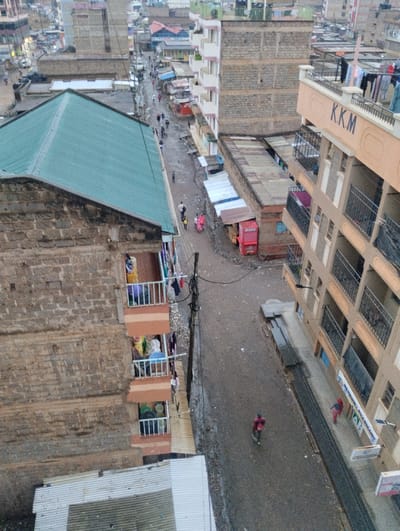 An aerial view depicts a densely populated urban neighborhood. Buildings of varying heights and construction styles line a narrow, wet street where people walk. Laundry hangs from balconies. Small shops are visible along the street, including a pharmacy. Overall, the scene conveys a sense of urban life in a low-income area.