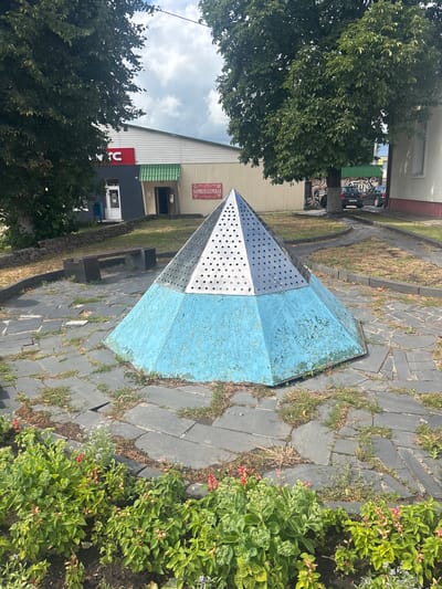 In the city of Капыльскі сельскі Савет, Belarus, a pyramid-shaped structure stands prominently in an outdoor setting. The base of the pyramid is painted a peeling, bright blue, while the upper section is made of perforated silver metal. It is situated on a paved area made of dark, irregular stone slabs, with patches of grass growing between them. Surrounding the paved area is a strip of greenery, featuring lush plants with red and pink flowers in the foreground. In the background, a building with a white and beige facade is visible, featuring a red sign with white lettering that reads "KFC". To the right of the KFC sign, a green awning extends over an entrance. Another building, painted beige, is to the right of the KFC building. A dark-colored car is parked in front of the beige building. Two dark concrete benches are placed behind the pyramid structure. The scene is framed by large trees with dense green foliage, suggesting a daytime setting with a partially cloudy sky.
