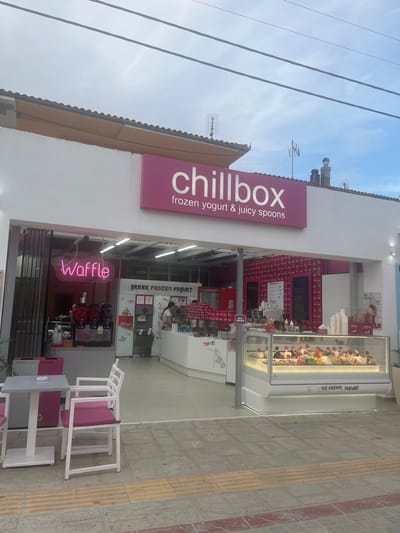 The image shows the exterior of a frozen yogurt and ice cream shop called "chillbox" located in Ofrynio Beach, Greece. The shop has a large pink sign with the name "chillbox" and the tagline "frozen yogurt & juicy spoons." In front of the shop, there is a display case filled with various flavors of ice cream. To the left, a neon sign spells out "Waffle" in pink. There are outdoor tables and chairs in front of the shop. The shop appears to be open for business, with staff visible inside. The environment is an outdoor commercial area with a paved walkway in front.