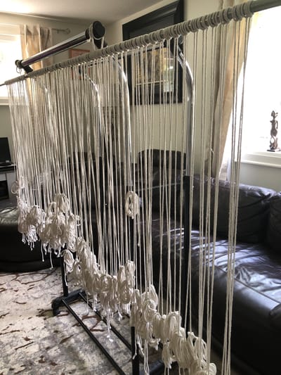 The image depicts a macrame setup in a residential living room. A series of white ropes are suspended from a horizontal bar, which is supported by a black metal stand. The ropes hang vertically, some with coiled knots near the bottom. In the background, a large brown leather sofa is visible, along with a patterned rug on the floor and a window with sheer curtains. The scene appears to be indoors, possibly in a home in Barrie, Canada, given the context of the request, although no specific identifying markers of the city are present in the image itself.