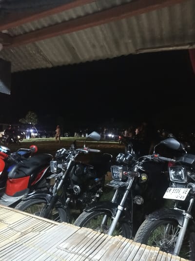 The image captures a nighttime gathering, viewed from beneath a corrugated metal roof. In the immediate foreground, a light-colored wooden or bamboo surface provides a base. Several motorcycles are parked in the mid-foreground, including a red and black scooter on the left, and at least three dark-colored standard motorcycles, some featuring square or rectangular LED headlights.
On the far right motorcycle, a white license plate reads "DR 5506 VE" with "12-23" below it, indicating an expiration date. A license plate on an adjacent motorcycle to its left partially shows "DL 4210". These license plate codes are characteristic of Indonesia, aligning with the provided location context of Kecamatan Pujut.
Beyond the parked motorcycles, an open dirt or grassy area is populated with numerous people standing and interacting in groups. One person stands somewhat centrally in the midground. Further in the distance, more figures and vehicle lights are visible against the dark night sky, suggesting a larger crowd or additional activities. The scene depicts a casual social event or meet-up, possibly for motorcycle enthusiasts, taking place outdoors or in a semi-sheltered space.