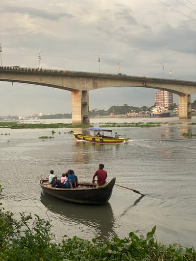 In Tarabo, Bangladesh, under a cloudy sky, a wide, murky river flows beneath a large, concrete bridge. The bridge has several supporting pillars and is elevated, with traffic visible on its surface, including what appears to be a car and people walking. Along the bridge's railing, there are streetlights and advertisements.
On the river, two boats are visible. In the foreground, a small, dark wooden rowboat contains four people: a man in a red shirt with a patterned waistcloth, who is rowing with an oar; a woman wearing a blue shirt and dark pants; a child in a pink shirt; and another person, possibly a child, in a light-colored shirt. This boat is positioned near the riverbank, which is densely covered with green aquatic plants and vegetation.
Further into the river, a larger, yellow and red boat with a blue canopy is moving away from the viewer, leaving a wake. This boat appears to be carrying several passengers, some of whom are visible under the canopy. In the background, a city skyline rises, featuring buildings and larger ships docked or anchored in the water. The overall scene depicts a busy waterway with both recreational and transport activities occurring under a somewhat overcast sky.