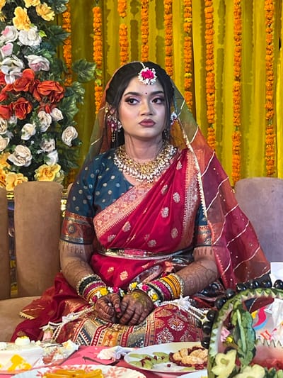 An adult female is depicted seated on a light brown upholstered sofa in Savar, Bangladesh. She is positioned against a yellow fabric backdrop adorned with multiple vertical garlands of orange marigold flowers. To her left, a large floral arrangement featuring white, orange, and yellow roses with green foliage is visible.
The woman wears elaborate traditional attire, including a deep red sari or lehenga with extensive gold embroidery and a teal blouse patterned with gold polka dots. A sheer red dupatta with gold trim is draped over her head and shoulders. Her adornments include a large, multi-layered gold and jeweled necklace, matching earrings, and a forehead ornament (maang tikka) with a pink floral accent. Multiple colorful bangles are visible on her wrists, which also display intricate henna designs extending up her forearms. Floral wrist decorations are present on both hands, which are clasped in her lap. Her hair is styled, and her face is made up, with a neutral expression and gaze directed slightly to the viewer's right.
In the foreground, a table is positioned in front of the woman, displaying several items. These include plates with sliced oranges or mandarins and scattered pomegranate seeds. An elaborately carved watermelon, possibly in the shape of a basket and filled with grapes and other fruits, is also present. A white cake with yellow accents is visible on a platter to the far left. A string of white pearls or beads lies near her hands.