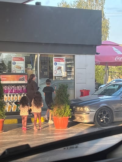 The image depicts a convenience store at a gas station in Tsaratsovo, Bulgaria, during daylight hours. The building is a modern, dark grey structure with large glass storefront windows. Inside the windows, multiple promotional posters with Cyrillic text "СПЕЦИАЛНО ПРЕДЛОЖЕНИЕ" (Special Offer) are visible, featuring items like a pastry. Shelves behind the glass display various products, including automotive oils branded "MEGATRON" in yellow and white containers.
An adult female with dark hair tied back, wearing dark clothing, stands near the store entrance, looking towards the right. Ahead of her, a boy in a dark t-shirt and shorts faces away from the camera, holding a blue bottle in his right hand. Closer to the foreground, two young girls, both wearing light patterned tops and maroon leggings, are seen from behind, walking towards the store entrance. The girl on the left wears pink shoes, and the girl on the right wears light pink shoes.
A grey BMW sedan, equipped with black multi-spoke aftermarket wheels, is parked to the right of the store entrance, with its front headlights visible. Several potted green plants in red plastic pots are placed around the entrance area and near the vehicle. Yellow concrete parking barriers are present on the paved ground. A red outdoor receptacle, possibly a bin or ashtray stand, is positioned next to the building on the right. A pink umbrella, branded "Coca-Cola," is visible on the far right, partially obscured by the building. In the background, green trees and a clear blue sky are evident. The group of four individuals appears to be approaching the convenience store.
