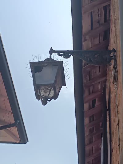 An old, rusted street lamp hangs from a decorative metal bracket attached to a textured orange wall. The lamp has broken glass panes and is covered in what appear to be bird deterrent spikes. To the left, a portion of a dark-colored awning or roof edge is visible against a pale blue sky. The architecture suggests an outdoor setting in an Italian city like Casalmaggiore, Italy.