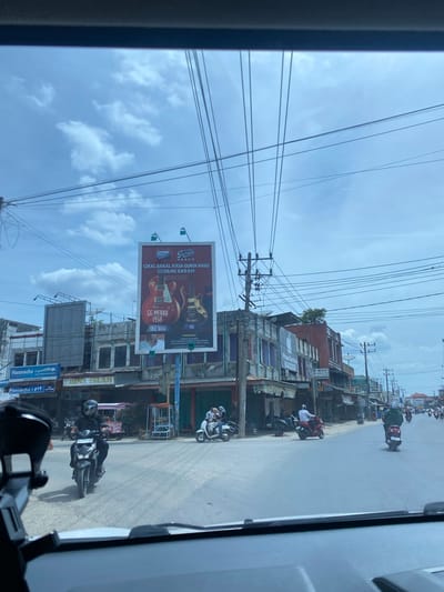 This media file captures a street scene in Meunasahpantonlabu, Indonesia. The image is dominated by a network of overhead power lines against a bright, cloudy sky. The perspective suggests the photo was taken from inside a vehicle, likely a car, looking out.

On the right side of the road, several buildings are visible, some appearing to be shops or businesses, with signs in Indonesian. One prominent billboard advertises "GUDANG GARAM MERAH" with images of guitars and the text "CIKAL BAKAL RASA GURIH KHAS" and dates "1958" and "1982." Another sign reads "Hannochs JAYA ELECTRONIC" and "BINA ISLAH."

The street is active with motorcycle traffic. Several individuals are seen riding motorcycles, some wearing helmets. The lighting suggests it is daytime, possibly late morning or early afternoon. The weather appears to be a mix of sun and clouds, with a few distinct cloud formations visible in the sky. The overall impression is of a bustling urban or semi-urban environment.

A small sign also mentions "REZA SERVICE HP," indicating a phone repair service is likely located nearby. The image is oriented vertically, with the sky occupying the upper portion and the street and buildings below.