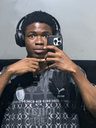 A young man takes a selfie with his black smartphone in what appears to be an indoor setting. He wears black headphones and a dark, patterned shirt with a Puma logo and a badge resembling a sports club emblem. He is touching his face with the hand holding the phone. The background consists of a light gray wall and a black vertical surface. The location is Ogwashi-Uku, Nigeria.