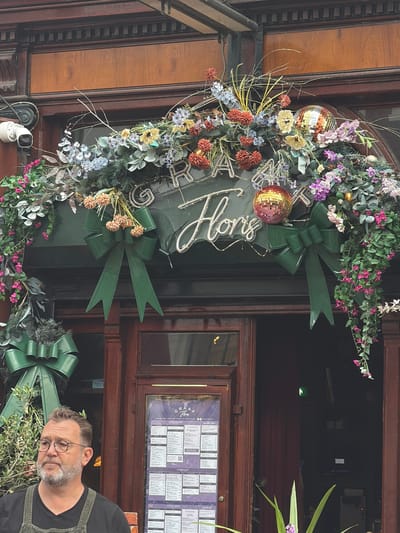 This image captures the decorated entrance of "GRAAF Floris" in Utrecht, Netherlands, with a man standing in the foreground. The entrance features a dark brown, ornate wooden frame, possibly a historical building style.
Dominating the upper section is an elaborate archway of artificial floral and foliage decorations. This includes a variety of flowers in shades of blue, purple, pink, orange/red, and yellow, interspersed with green leaves, pine needles, and bare twigs. Integrated into this display are several small, reflective disco balls and delicate fairy lights. The establishment's name, "GRAAF Floris," is prominently displayed: "GRAAF" in individual silver block letters, and "Floris" in an illuminated, white or silver script font. Below this, two large, dark green decorative bows are affixed to the door frame on either side. A third, smaller green bow is visible lower on the left.
To the upper left of the entrance, a white security camera (CCTV) is mounted on the wall. The entrance consists of dark brown wooden doors with glass panels. On the right door panel, a large purple/dark blue menu board is displayed, titled "GRAAF Floris" and listing "LUNCHKAART" (Lunch Menu) and "DINERKAART" (Dinner Menu) in Dutch, with some English translations. The right door is partially open, revealing a dark interior with what appears to be a reddish curtain and a faint green exit sign.
In the lower-left foreground, a middle-aged man is partially visible. He has short, light brown/grey hair, a neatly trimmed beard, and wears round-rimmed glasses. He is dressed in a dark t-shirt underneath dark overalls or dungarees. His gaze is directed to his left, away from the camera, with a neutral expression. To his left, a potted plant with green leaves and small olive-like branches is visible. In the lower-right corner, additional green foliage and a single purple flower from another plant are seen. The scene is illuminated by natural daylight, suggesting an overcast day or shaded area.