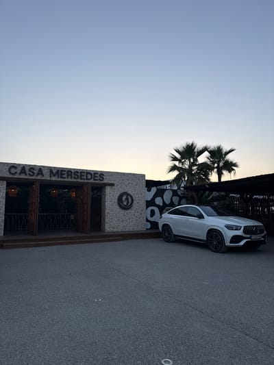 A white SUV is parked on a paved surface next to a building with "CASA MERSEDES" written vertically on its facade. The building has a stone exterior and features several levels of windows with wooden accents. Above the windows, a circular emblem is visible. Two palm trees are situated behind and to the left of the building, with their fronds silhouetted against a light-colored sky, suggesting it is either early morning or late evening. The sky is clear and gradient, transitioning from a light blue to white. The overall scene appears to be an outdoor setting, possibly a restaurant or establishment, in a warm climate. The license plate on the SUV is partially visible and appears to read "TJ2-M-599". The image is oriented vertically, with the sky at the top.