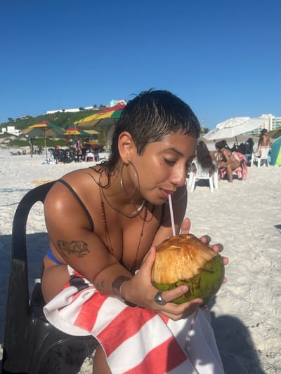 At the beach, a woman sips from a coconut with a straw. She wears large hoop earrings and several necklaces. A tattoo is visible on her upper arm. A striped towel rests on her lap. The background reveals sunbathers, umbrellas, and beachside buildings under a clear sky. The photo is rotated sideways.