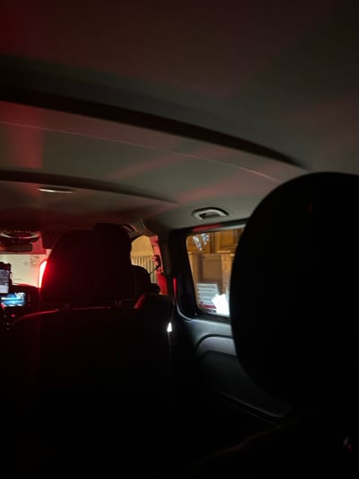 An angled view from inside a car shows the passenger side. A sun visor is partially lowered. A rear window reveals a lit building exterior. Car seats and other vehicle components are partially visible in the dimly lit interior. The passenger side is a light gray. A handle and indentation are on the passenger side panel. A red glow illuminates the back of the vehicle.