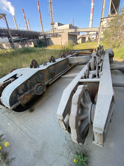 The image depicts an industrial site with heavy machinery and infrastructure. In the foreground, two large, metallic structures, possibly parts of a conveyor system or rail car, are visible on a concrete surface. One structure has a caterpillar-like track, while the other features large wheels and a complex arrangement of gears and rollers. The background shows a vast industrial complex with numerous smokestacks emitting white and grey plumes, along with elevated walkways and buildings. Greenery, including tall grass and some yellow wildflowers, grows around the industrial elements, suggesting a period of disuse or natural reclamation. The sky is clear and blue. The context provided indicates this scene is located in the city of Police, Poland. There are no people visible in the image.