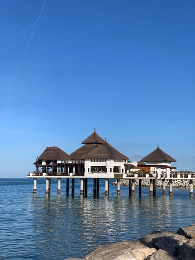 The image captures an overwater structure in M'diq, Morocco, under a clear, bright blue sky with a faint white contrail diagonally traversing the upper left quadrant. The central subject is a series of three connected buildings constructed on numerous cylindrical stilts over a calm, blue-green body of water. Each building features a white facade and a prominent, multi-tiered, conical roof made of dark brown thatched material. The largest building is centrally located, flanked by two smaller, similarly styled structures. Visible details include dark-framed windows, balconies with dark railings, and what appear to be outdoor seating arrangements on the terraces, particularly on the leftmost building. A pier-like extension, also supported by stilts, connects the rightmost building to a man-made breakwater composed of large, grey-white rocks. In the immediate foreground, to the lower right, additional large, irregular grey rocks indicate a shoreline or bank. Small white birds are visible perched on the roofs of the leftmost building and flying above the water near it. The water surface displays minor ripples and reflects the sky and structures. No human figures are discernible within the frame.