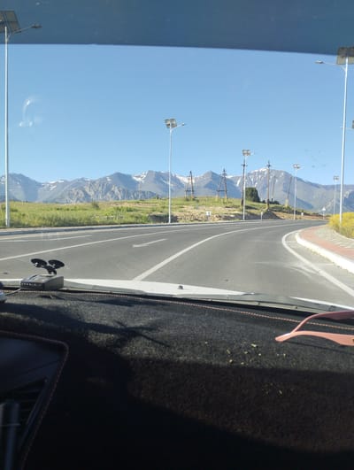 The image shows a view from the interior of a car looking out at a road. The road is paved and has white lane markings. There are solar-powered streetlights along the side of the road. In the distance, there are mountains with snow-capped peaks. The sky is clear and blue. There is a dashboard in the foreground of the car, with a radar detector mounted on it. To the right of the radar detector, there are pink-rimmed sunglasses on the dashboard. The image appears to have been taken in a rural or mountainous area. There is no indication that the image was taken in Myk, Uzbekistan.