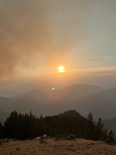 The image depicts a sunset over a mountainous landscape, with the sun appearing as a bright orb in a hazy, orange-tinted sky. Layers of silhouetted mountains recede into the distance, creating a sense of depth. In the foreground, a dense forest of dark evergreen trees stands on a hillside, with dry, golden grass covering the ground. The scene is natural and serene, with no signs of human activity or urban presence, therefore it cannot be placed in San Juan de Plan.