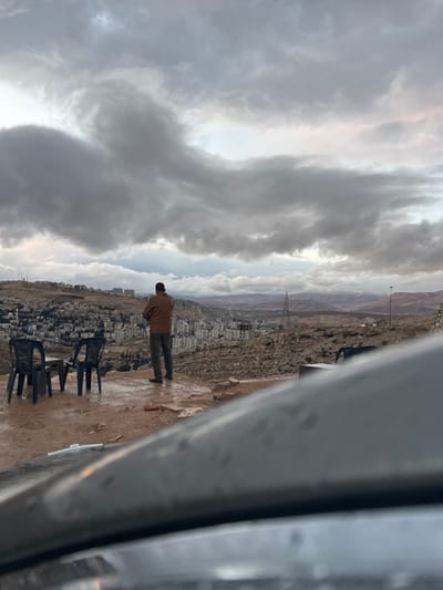 A wide-angle image depicts an elevated view over the city of Dummar Municipality, Syria, under a heavily overcast sky. In the mid-ground, a solitary adult male, viewed from behind, stands on a wet, reddish-brown rocky outcrop. He wears a brown jacket and dark trousers, with his hands clasped in front of him, facing the sprawling cityscape below. To his left, two dark-colored plastic chairs and a small dark table are arranged on the wet ground.

The city of Dummar extends across the middle ground, comprising numerous multi-story residential buildings constructed into hilly terrain. Many building lights are illuminated, suggesting late day or evening conditions. Beyond the city, the landscape consists of rugged, rocky hills. A tall, metallic power transmission tower stands on a distant hillside to the right of the city. Further in the background, snow-capped mountains are visible on the horizon. The sky is dominated by thick, dark grey clouds, with some lighter patches visible closer to the horizon. The immediate foreground shows the wet, dark surface of a vehicle's hood, indicating the photograph was taken from inside a car or from a vehicle's perspective. The ground throughout the visible rocky areas appears wet, consistent with recent precipitation.