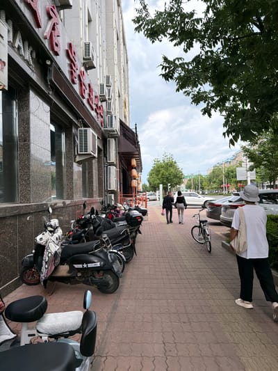 The image depicts a street scene in Hunchun, China. In the foreground, several parked scooters and electric bikes line the sidewalk in front of a building with a granite facade and red Chinese characters on its exterior. The building also features numerous air conditioning units mounted on its wall. Further down the sidewalk, two individuals are walking away from the camera, followed by a person wearing a hat and carrying a tote bag. A bicycle is parked on the sidewalk near them. The street is lined with trees, and cars are visible parked along the opposite side of the street. The sky is overcast.