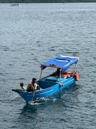 Two dark-skinned men navigate a narrow, bright blue boat on rippling water off Rinca Village, Indonesia. One man, wearing a white shirt, sits facing forward, rowing with an oar. Behind him sits another person, with dark hair, whose face is partially visible. The boat is covered with a blue-and-white awning, with red gear visible toward the front. The water is textured with small waves, reflecting sunlight. In the distance, on the left side, is a land mass covered in green vegetation and a smaller boat.