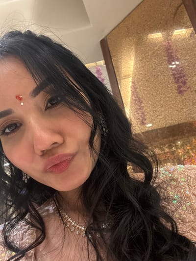 A woman with dark, wavy hair is angled towards the camera. She has fair skin, dark eyes, and a red bindi on her forehead. She wears a diamond necklace and earrings. The backdrop is a textured surface with golden and pink tones. Her outfit has sequins and intricate detailing. The lighting is soft and warm.