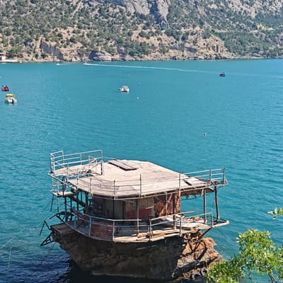 This media file depicts a rustic, weathered wooden structure perched atop a rocky outcrop in the bright, turquoise waters of a bay. The structure itself appears to be a multi-level platform with a small, enclosed cabin-like section at its base. It features a wooden deck, metal railings that are somewhat rusted, and exposed wooden beams and supports. There are benches visible on the deck. The rock formation it sits on is rough and textured, partially submerged in the water. Ropes are attached to the rock and the structure, securing it.
In the background, a rugged, mountainous coastline covered in dense green vegetation slopes down to the water. Several small boats are visible in the bay, some moving and creating wakes, suggesting a leisurely or recreational activity. One boat is closer to the foreground on the left, and others are further out towards the coastline. The water is calm and clear, reflecting the bright sunlight.
The scene is illuminated by bright daylight, indicating a sunny day, likely mid-morning or afternoon. The clear water and vibrant colors suggest pleasant weather conditions. The overall impression is one of a somewhat isolated, perhaps abandoned or derelict, man-made structure in a beautiful natural setting. There is no visible text in the image. The location context provided is Novyi Svet, Ukraine.