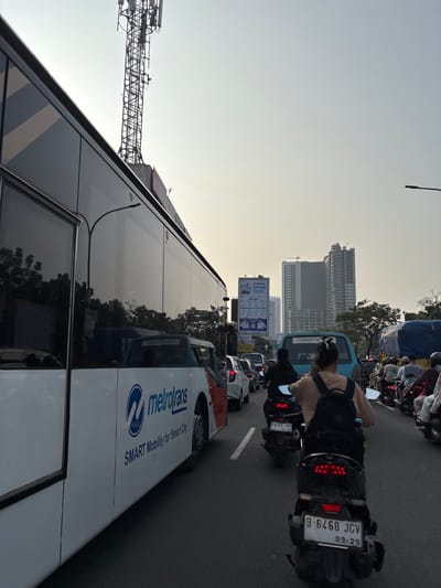The image depicts a congested urban street scene in South Tangerang, Indonesia, likely during daylight hours, possibly late afternoon due to the warm, diffused light. A large white bus, branded "metrotans SMART Mobility for Smart City," dominates the left side of the frame, its reflective windows mirroring the surrounding environment and traffic. In the distance, several modern high-rise buildings, one appearing to be under construction, punctuate the skyline.
The street is filled with a variety of vehicles, primarily motorcycles and cars, indicating significant traffic congestion. Several motorcyclists, some with passengers, are visible, navigating through the traffic. One motorcyclist in the foreground is wearing a light brown hooded jacket and a black backpack. The license plate of this motorcycle is clearly visible: B-6468 JGV, with an expiry date of 09-29. Another motorcycle behind it has the license plate B-6326 JCD. A light blue van is also present in the traffic lane.
A tall telecommunications tower is a prominent vertical element in the background, on the left side of the image. Billboard advertisements are also visible, including one with text in Indonesian, promoting something related to "JADWAL SPMB." Trees are scattered throughout the scene, and a street lamp is visible over the bus. The road surface is asphalt, marked with white lane lines. The overall impression is one of a busy, bustling city environment experiencing heavy traffic.