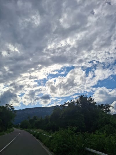 The image captures a scenic mountainous landscape in Lyutibrod, Bulgaria, under a dramatic sky. The perspective is from a slightly elevated viewpoint, looking down a winding asphalt road.
The sky, occupying a significant portion of the frame, features a striking interplay of bright white and grey clouds, interspersed with patches of clear blue sky. Sunlight is visible breaking through the clouds in several areas, creating a dynamic and atmospheric lighting. The cloud patterns suggest shifting weather conditions, possibly after a recent rain or during a period of changing weather.
On the right side of the photograph, a paved road with a distinct white solid line curves gently into the distance. It is flanked by dense, lush green vegetation, including tall trees, various bushes, and ferns, indicating a rich natural environment. A white guardrail can be seen partially along the edge of the road, amidst the greenery.
In the background, a large, dark mountain dominates the horizon, its imposing form a prominent feature against the sky. The mountain appears verdant with deep green foliage.
The scene depicts a tranquil, natural setting, devoid of people or animals. There is no visible text in the image. The lighting indicates it is daytime, possibly mid-morning or late afternoon, given the dramatic interplay of light and shadow on the clouds and landscape.