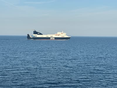 A DFDS ferry, identified by the prominent branding on its hull and superstructure, sails across a calm blue ocean under a clear sky with faint clouds. The ferry is dark blue with a white upper deck and has a single funnel emitting a plume of smoke. The vessel appears to be moving from left to right in the frame. The water is rippled, indicating a gentle breeze. The horizon is visible where the sea meets the sky, with the sky occupying the upper portion of the image. The scene suggests an open sea environment, likely a shipping route.
