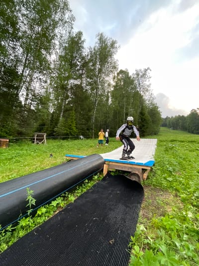 An individual wearing a helmet, goggles, and protective gear is shown on a makeshift ramp constructed from wood and a textured mat. The person is on a snowboard, appearing to be in motion. Behind them, two other individuals stand on a grassy slope, one wearing a yellow shirt and the other in white. The scene is set outdoors, surrounded by lush green grass and tall trees, suggesting a natural environment. In the background, a ski lift or similar structure is visible on a distant hill, indicating the location might be a ski resort or a recreational area. The sky is overcast with clouds. The overall impression is of an outdoor activity, possibly training or practice, taking place in a summer or early autumn season. This image depicts a recreational area in Divnogorsk, Russia, potentially a ski resort or a park, given the presence of a ski lift and the mountainous terrain.