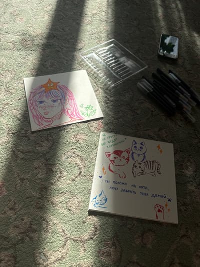 The image captures an indoor scene in Uryupinsk, Russia, with direct sunlight casting sharp shadows across a light green and gray patterned carpet. The angle of the sunbeams suggests either morning or late afternoon.
The primary subjects are two pieces of paper displaying vibrant drawings. The larger drawing features three stylized cats: one blue, one red, and one striped brown, alongside various decorative elements like hearts, stars, and paw prints. Handwritten Russian text accompanies the illustrations. On the left, it reads, "НЕ ГРУСТИ ВСЁ БУДЕТ КЛУБНИЧНО" (Don't be sad, everything will be strawberry). On the right, it says, "- ТЫ ПОХОЖА НА КОТА, ХОЧУ ЗАБРАТЬ ТЕБЯ ДОМОЙ." (- You are like a cat, I want to take you home.).
The smaller drawing, situated to the lower left, depicts an anime-style character with flowing pink and blue hair and large blue eyes, seemingly smiling. A small yellow star sticker is affixed to the character's hair, and faint green clover or leaf designs are drawn in the top corner.
Around the drawings are art supplies: a collection of black markers or pens with colored caps, and a clear plastic container, possibly for holding pens. In the upper left corner, a small, silver-framed mirrored object reflects a blurry image of green foliage. The scene evokes a feeling of creative leisure and conveys comforting and affectionate sentiments through the illustrations and Russian text.