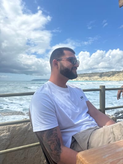 An adult male is seated in profile, looking right, against a coastal backdrop identified as Episkopi Lemesou, United Kingdom. He possesses short dark hair, a beard, and wears dark sunglasses. His attire consists of a white short-sleeved t-shirt featuring a dark "Alpha Industries" logo on the left chest and khaki-colored shorts. A detailed tattoo, appearing to depict a face within an ornate design, covers his left forearm/bicep. His left arm rests on a light-colored wooden table in the foreground. Behind him, a wooden railing separates the seating area from a choppy blue-green sea with visible white-capped waves breaking on a rocky shore. A light-colored, rugged cliff or headland is visible across the bay in the middle distance. The sky above is clear blue with scattered white cumulus clouds. A second person's hand is partially visible on the far right, resting on the wooden railing.