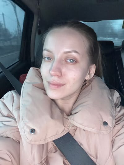 A female individual is depicted seated within a vehicle's passenger compartment, specifically in Spas-Zaulok, Russia. She is secured by a dark-colored seatbelt extending across her chest and left shoulder. The individual has fair skin, light blue-grey eyes, and light brown hair pulled back from her face. She wears a light beige or pale pink quilted puffer jacket, featuring visible metallic snap fasteners on the collar. Small, geometric, possibly gold-toned earrings are present on both ears. Her expression is neutral with a slight upward curve to her lips, and she maintains direct eye contact with the viewer. The vehicle's interior is dark. Through the left-side window, an indistinct exterior environment is visible under dim, overcast lighting, characterized by blurred roadside elements and sparse vegetation. A small, dark hanging ornament is suspended near the upper frame of the left window. A dark headrest is partially discernible behind the individual.