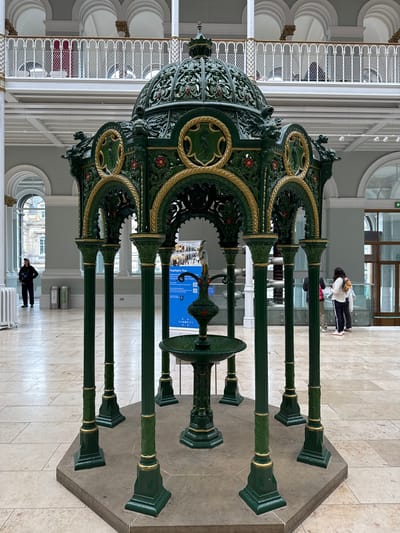 This is a medium-shot, eye-level, full-color photograph of a tall, ornate, dark green and gold fountain in the Great Hall of the National Museum of Scotland, located in the city of Edinburgh, United Kingdom. The fountain is centrally located and occupies the foreground of the image. It features a domed canopy adorned with intricate metalwork and gold accents, supported by several slender, fluted columns. Decorative circular panels depicting wildlife are integrated into the structure. A smaller basin with a bird-like sculpture sits at the base of the fountain.
The background reveals the architecture of the Great Hall, characterized by a multi-level interior with pale tiled floors, light grey walls, and large arched windows. A white balustrade with gold trim runs along an upper level, where several indistinct figures can be seen. In the middle ground, a blue sign with text and a QR code is placed to the right of the fountain, indicating a "Highlights Tour." Two silver trash receptacles are visible in the left mid-ground.
In the far left, a person wearing dark clothing and a cap stands facing away from the camera. In the middle ground, slightly to the right of the fountain, a group of three people, two women and one man, are looking at a piece of paper. The woman on the left is wearing a white top and dark pants, while the woman on the right has a yellow backpack. The man is wearing a dark top and light-colored pants. Further to the right, in the background, two people are visible in front of a large glass door, one wearing a white top and the other a darker top. The overall environment suggests an indoor public space, likely a museum or gallery, with a bright, diffused lighting.
