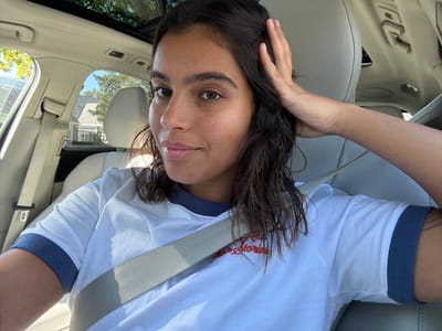 A young adult female is depicted in a medium close-up, seated in the front passenger seat of a light-colored vehicle, secured by a light gray seatbelt. She has medium-length dark hair, styled to the side, and is looking directly at the camera with a subtle smile. Her right hand is raised, with fingers touching the hair above her ear. She is wearing a white short-sleeved ringer t-shirt with blue trim on the collar and sleeves. Red embroidery, partially obscured, is visible on the front of her shirt. The car's interior features light-colored upholstery, likely leather, and a panoramic sunroof is visible overhead, allowing bright sunlight into the cabin. Through the vehicle's windows, a residential scene is visible under a clear blue sky, featuring a large house with light siding and a dark roof, surrounded by green foliage, consistent with a suburban environment in Princeton, United States.