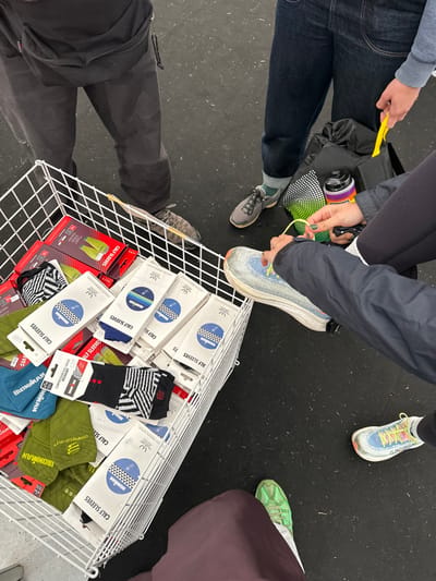 This image depicts an overhead view of a scene located in Weymouth, United Kingdom, focusing on the lower extremities of at least four individuals surrounding a white wire retail display bin. The bin is filled with packaged athletic products, predominantly "CALF SLEEVES" and "COMPRESSION SOCKS RECOVERY" displaying "Ironman" branding in various colors including white, blue, black, and green.
To the upper-left, a person is visible wearing dark grey trousers and dark grey athletic shoes. To the upper-right, an individual wears dark blue jeans, green socks, and grey athletic shoes, holding a yellow packet or wrapper in their right hand. Centered to the right, another person in dark athletic wear and a dark jacket is actively tying the light blue shoelace of a prominent white and light blue athletic shoe. A second, white and multi-colored athletic shoe with yellow laces is visible near this person's lower right foot. Adjacent to them on the dark floor is a black drawstring bag, holding a multi-colored water bottle with a black cap. To the lower-left, a fourth person wears a dark maroon lower garment and a bright green athletic shoe. The floor is a uniform dark surface, likely black or dark grey, scattered with fine debris.