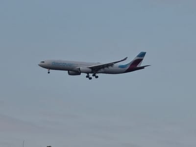 An Airbus A330 aircraft, operated by Discover Airlines, is captured mid-flight against a pale blue, overcast sky. The aircraft is predominantly white with blue lettering that reads "discover" along the fuselage. The tail fin displays a distinctive blue and maroon geometric pattern. The landing gear is extended, indicating the aircraft is either preparing for landing or has just taken off. The scene is consistent with the flight path over Kelsterbach, Germany, an area near Frankfurt Airport, a major hub for international air traffic.