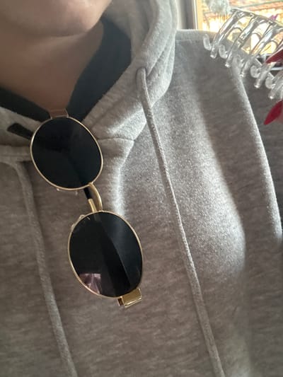 The image shows a pair of gold-framed, round sunglasses resting on a grey hooded sweatshirt. Part of a clear plastic hair clip is visible in the upper left corner. A sliver of skin is visible to the left. The location is not identifiable as Lehchevo, Bulgaria.